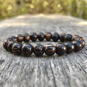 May include: A beaded bracelet featuring dark brown wooden beads with a striped pattern. The bracelet is resting on a weathered wooden surface, with a blurred background of green foliage.