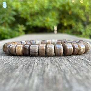 以下が含まれることがあります： 茶色とベージュの色合いの、磨かれた長方形の木製ビーズで作られたブレスレット。 ブレスレットは、ぼやけた緑色の背景を持つ、風化した木製の表面に置かれています。