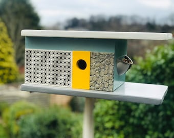 May include: A modern birdhouse with a geometric design. The birdhouse features a light blue body, a yellow accent, and a textured stone-like panel. It has a small metal feeder and a perch. The roof is white and gray.
