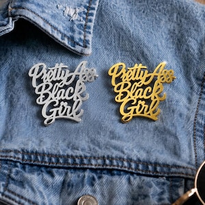 May include: Two script-style lapel pins attached to a blue denim jacket. One pin is silver-toned and the other is gold-toned. Both pins feature the text "Pretty Ass Black Girl" in a cursive, stacked font design with a reflective, metallic finish.