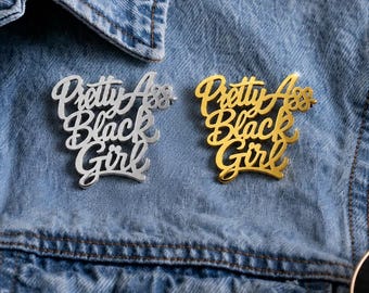 May include: Two script-style lapel pins attached to a blue denim jacket. One pin is silver-toned and the other is gold-toned. Both pins feature the text "Pretty Ass Black Girl" in a cursive, stacked font design with a reflective, metallic finish.
