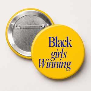 May include: Two yellow round buttons. One button is face up, displaying the text "Black girls Winning" in blue. The other button is face down, showing the silver pin back. The buttons are likely for clothing or accessories.