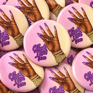 May include: A collection of round buttons with a hand illustration. The hand is brown with long purple nails, a gold watch, and jewelry. The text "Clock That Tea" is in purple script on a pink background.