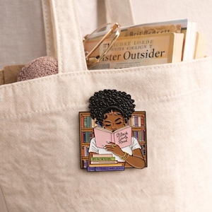 Pode incluir: Uma sacola creme com livros e um alfinete. O alfinete apresenta uma mulher negra lendo um livro rosa com o texto "Black Girls Read". O alfinete est&aacute; em um fundo de estante. A sacola cont&eacute;m livros e um pequeno objeto rosa redondo.