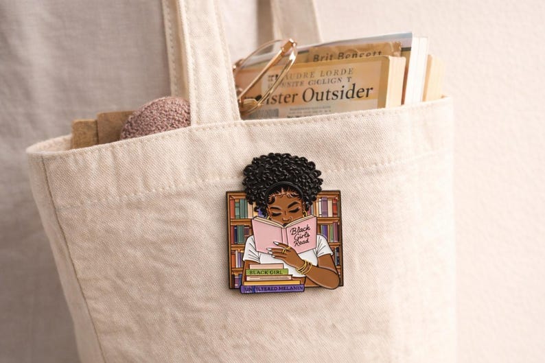 Pode incluir: Uma sacola creme com livros e um alfinete. O alfinete apresenta uma mulher negra lendo um livro rosa com o texto "Black Girls Read". O alfinete est&aacute; em um fundo de estante. A sacola cont&eacute;m livros e um pequeno objeto rosa redondo.