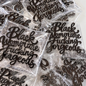 May include: A collection of black metal pins, each individually packaged in a clear plastic sleeve. The pins feature the cursive text Black Women are Fucking Gorgeous. Each pin has a metallic finish and a butterfly clutch backing visible through the plastic.