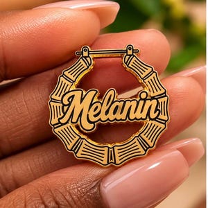 May include: A gold-toned circular hoop earring featuring a bamboo-inspired texture and a horizontal bar at the top. The center of the hoop displays the word Melanin in a bold, black, cursive script font with a gold-toned outline.