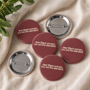 May include: A collection of burgundy-colored round buttons with the text "dear Black women, you are the standard." The buttons have a silver pin backing. The buttons are arranged on a beige fabric.