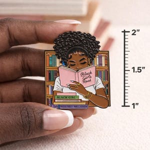 Pode incluir: Um alfinete de esmalte retangular com uma mulher negra lendo um livro rosa com o texto "Black Girls Read". O alfinete tem aproximadamente 5 cm de altura. A mulher est&aacute; em frente a uma estante.