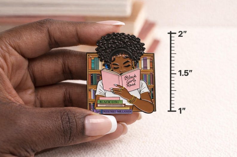 Pode incluir: Um alfinete de esmalte retangular com uma mulher negra lendo um livro rosa com o texto "Black Girls Read". O alfinete tem aproximadamente 5 cm de altura. A mulher est&aacute; em frente a uma estante.