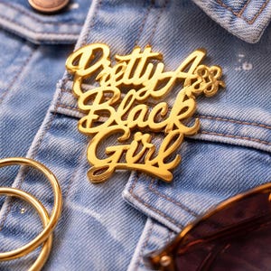 May include: A gold-colored pin with the words "Pretty Ass Black Girl" in a cursive font is pinned to a light blue denim jacket. Gold hoop earrings and sunglasses are also visible.