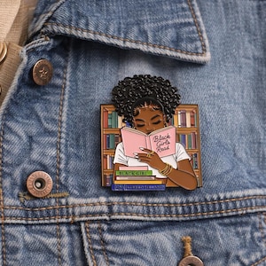 Pode incluir: Um alfinete de esmalte colorido com uma mulher negra lendo um livro com o texto "Black Girls Read". O alfinete est&aacute; colocado em uma jaqueta jeans, com uma estante e livros empilhados no fundo. Um acess&oacute;rio vibrante.