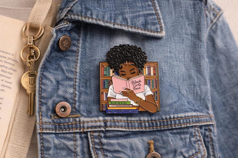 Pode incluir: Um alfinete de esmalte colorido com uma mulher negra lendo um livro com o texto "Black Girls Read". O alfinete est&aacute; colocado em uma jaqueta jeans, com uma estante e livros empilhados no fundo. Um acess&oacute;rio vibrante.