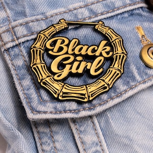 May include: A gold-colored pin with the words "Black Girl" in a stylish font, set within a bamboo-style hoop. The pin is displayed on a light blue denim jacket, showcasing its design and color contrast.
