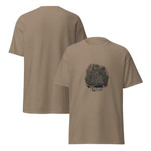 May include: A tan short-sleeved t-shirt featuring a dark, circular graphic on the front chest. The graphic depicts a detailed map illustration with the text VERDANSK printed in bold, capital letters at the bottom of the circular design.