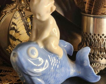 May include: A porcelain figurine featuring a white child figure sitting atop a light blue fish. The fish has a smooth, glossy finish with visible scale textures and a prominent eye. The child figure is depicted in a seated position on the back of the fish, with a smooth, matte-like white surface. The entire piece is crafted as a single, cohesive decorative object.