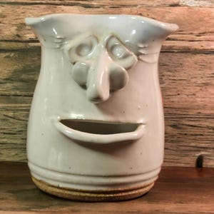 May include: White ceramic pitcher with a face sculpted on the front. The face has a large nose, wide eyes, and a wide mouth.