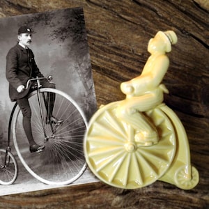 May include: A vintage photograph of a man riding a penny-farthing bicycle, next to a ceramic figurine of a person riding a similar bicycle. The figurine is painted in a pale yellow color.