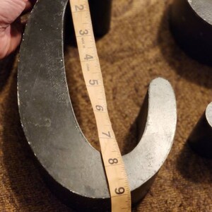 Puede incluir: Dos letras met&aacute;licas grandes, una con forma de "C" y la otra con forma de "U", se est&aacute;n midiendo con una cinta m&eacute;trica. Las letras est&aacute;n pintadas de negro y tienen un aspecto ligeramente desgastado y envejecido.