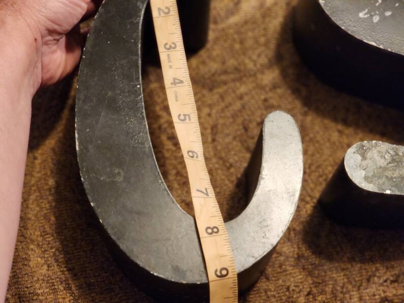 Puede incluir: Dos letras met&aacute;licas grandes, una con forma de "C" y la otra con forma de "U", se est&aacute;n midiendo con una cinta m&eacute;trica. Las letras est&aacute;n pintadas de negro y tienen un aspecto ligeramente desgastado y envejecido.