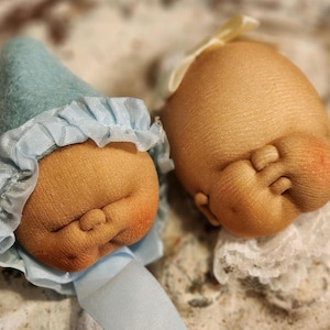 May include: Two handmade cloth dolls with closed eyes, one wearing a blue and white hat and the other wearing a white lace bonnet.