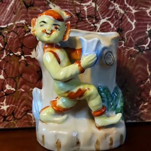 May include: A ceramic figurine of a yellow elf with orange stripes holding a blue flower. The elf is standing on a tree stump with green leaves. The figurine is a vintage style planter.