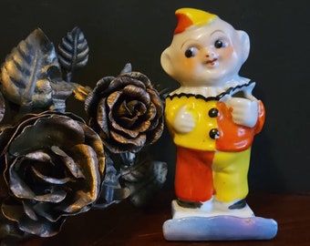 May include: A porcelain figurine of a clown with a yellow, red, and white costume. The clown is holding a small white object in its right hand. The figurine is standing on a blue and white base.