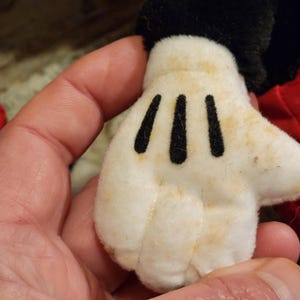 May include: A white plush glove with black stitching, resembling a cartoon character's hand. The glove has three black lines on the palm and is slightly stained with brown marks.
