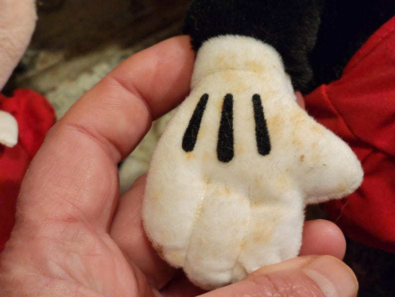May include: A white plush glove with black stitching, resembling a cartoon character's hand. The glove has three black lines on the palm and is slightly stained with brown marks.