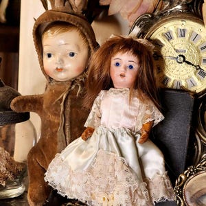 May include: Two antique dolls are featured in this image. One doll is dressed in a brown bunny suit, and the other doll is wearing a light pink dress with lace trim. The dolls have painted faces and are set against a vintage clock.