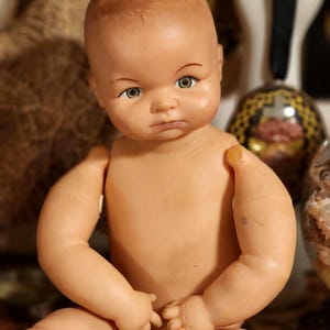 May include: A vintage doll with a smooth, light-toned plastic body, molded hair, and painted facial features including green eyes and arched eyebrows. The doll has articulated shoulders and is depicted in a seated position with a neutral expression.
