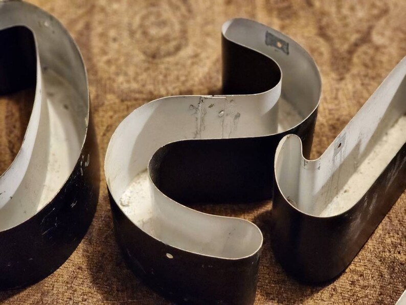 Puede incluir: Tres letras de metal negro, cada una con forma de letra 'C', est&aacute;n dispuestas en fila. Las letras son huecas y tienen un interior blanco. Las letras est&aacute;n sobre una superficie marr&oacute;n.