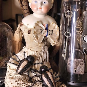 May include: A vintage porcelain doll with blonde hair, blue eyes, and rosy cheeks, wearing a cream-colored dress and a bow with the year "1957." Two decorative spiders rest on a lace doily. A glass cloche with keys is in the background.
