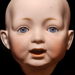 May include: A porcelain doll head featuring a smooth, pale complexion with rosy cheeks, bright blue glass eyes, painted light brown eyebrows, and an open mouth revealing small teeth. The doll has a bald, rounded head and prominent ears.