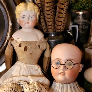 May include: Two antique porcelain dolls, one complete and one head only, with detailed facial features and vintage clothing. The complete doll has a blonde wig and a white dress. The head has round glasses and a ruffled collar. Decorative feathers are in the background.