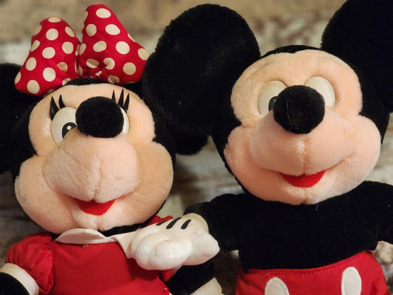 May include: A plush Mickey Mouse and Minnie Mouse toy set. Mickey is wearing red shorts and Minnie is wearing a red dress with a white collar and a red and white polka dot bow.