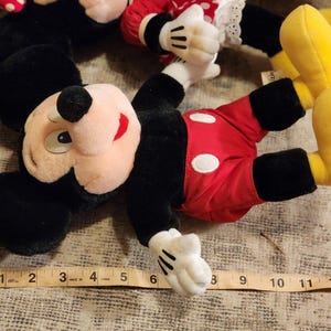 May include: A plush Mickey Mouse toy with black ears, a red and white polka dot shirt, and yellow shoes. The toy is lying on a brown surface with a measuring tape.