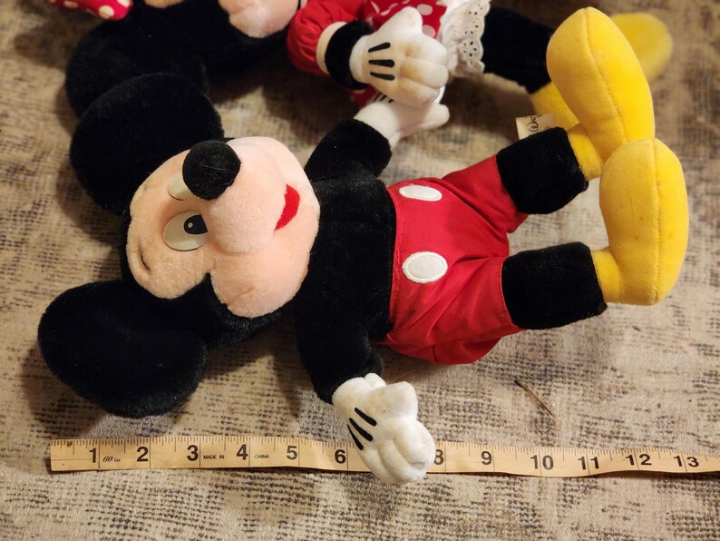May include: A plush Mickey Mouse toy with black ears, a red and white polka dot shirt, and yellow shoes. The toy is lying on a brown surface with a measuring tape.