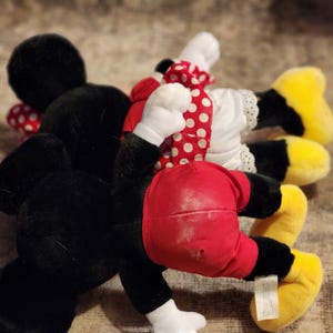 May include: A plush toy set featuring Mickey Mouse and Minnie Mouse. Mickey is wearing a black and red outfit, while Minnie is wearing a white dress with red polka dots and a red bow.