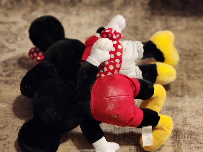 May include: A plush toy set featuring Mickey Mouse and Minnie Mouse. Mickey is wearing a black and red outfit, while Minnie is wearing a white dress with red polka dots and a red bow.