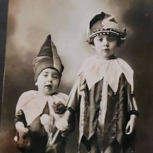 May include: Two children dressed as jesters, one holding a stuffed animal. The child on the left is wearing a pointed hat with red, white, and blue accents. The child on the right is wearing a jester hat with bells and a white collar.