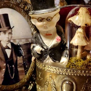 May include: A vintage framed photo of a man wearing a top hat and suit, next to a porcelain figurine of a person wearing a top hat and suit, and a glass dome containing dried mushrooms.