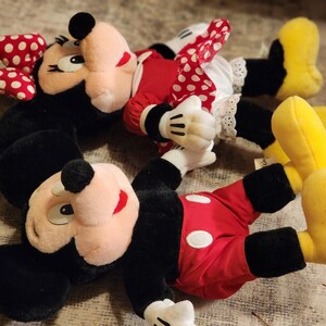 May include: Two plush toys of Mickey Mouse and Minnie Mouse. Mickey Mouse is wearing red shorts with a white circle and black shoes. Minnie Mouse is wearing a red and white polka dot dress with a white petticoat and yellow shoes.