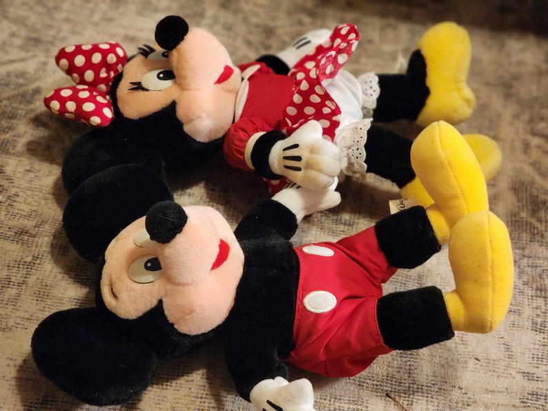 May include: Two plush toys of Mickey Mouse and Minnie Mouse. Mickey Mouse is wearing red shorts with a white circle and black shoes. Minnie Mouse is wearing a red and white polka dot dress with a white petticoat and yellow shoes.