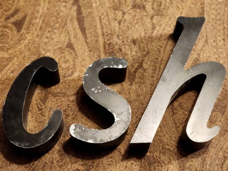 Puede incluir: Tres letras met&aacute;licas, C, S y H, en un acabado negro ligeramente desgastado. Las letras son estilizadas y tienen un aspecto vintage.