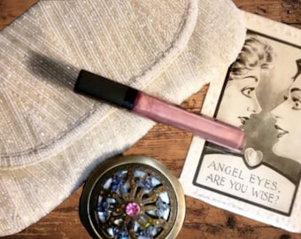 May include: An ivory beaded clutch, a pink lip gloss, and a vintage compact mirror with blue and pink accents are arranged on a wooden surface. A vintage-style card with the text "ANGEL EYES, ARE YOU WISE?" is also present.