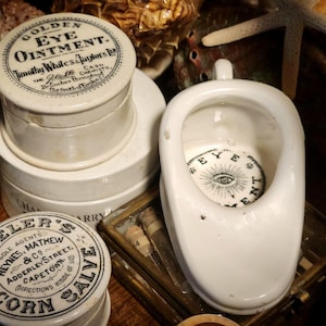 May include: An antique collection featuring vintage apothecary jars and a white porcelain urinal. The jars are labeled with text such as "Golden Eye Ointment" and "Corn Salve". The urinal has an eye design inside.