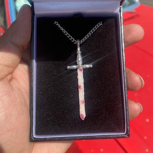 Könnte beinhalten: Eine silberne Schwertkette mit einem weißen und rosafarbenen Glitzerdesign. Die Kette befindet sich in einer schwarzen Samtbox.