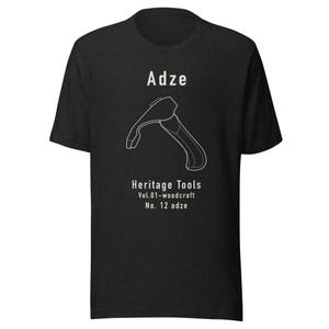 May include: Dark gray t-shirt featuring a white line drawing of an adze, a woodworking tool. The text "Adze" is above the drawing, with "Heritage Tools" and "Vol.01-woodcraft No. 12 adze" below. The shirt is a short-sleeved crew neck.