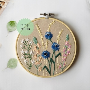 May include: A PDF pattern for an embroidery hoop featuring a floral design with blue, white, pink, and yellow flowers on a beige background. The text "PDF PATTERN" is in a green circle.